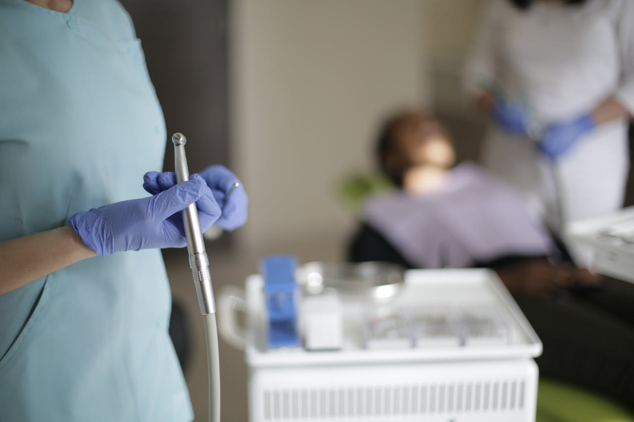 Unrecognizable dentist in uniform and gloves standing with dental drill in selective focus while blurred assistant preparing patient for treating teeth